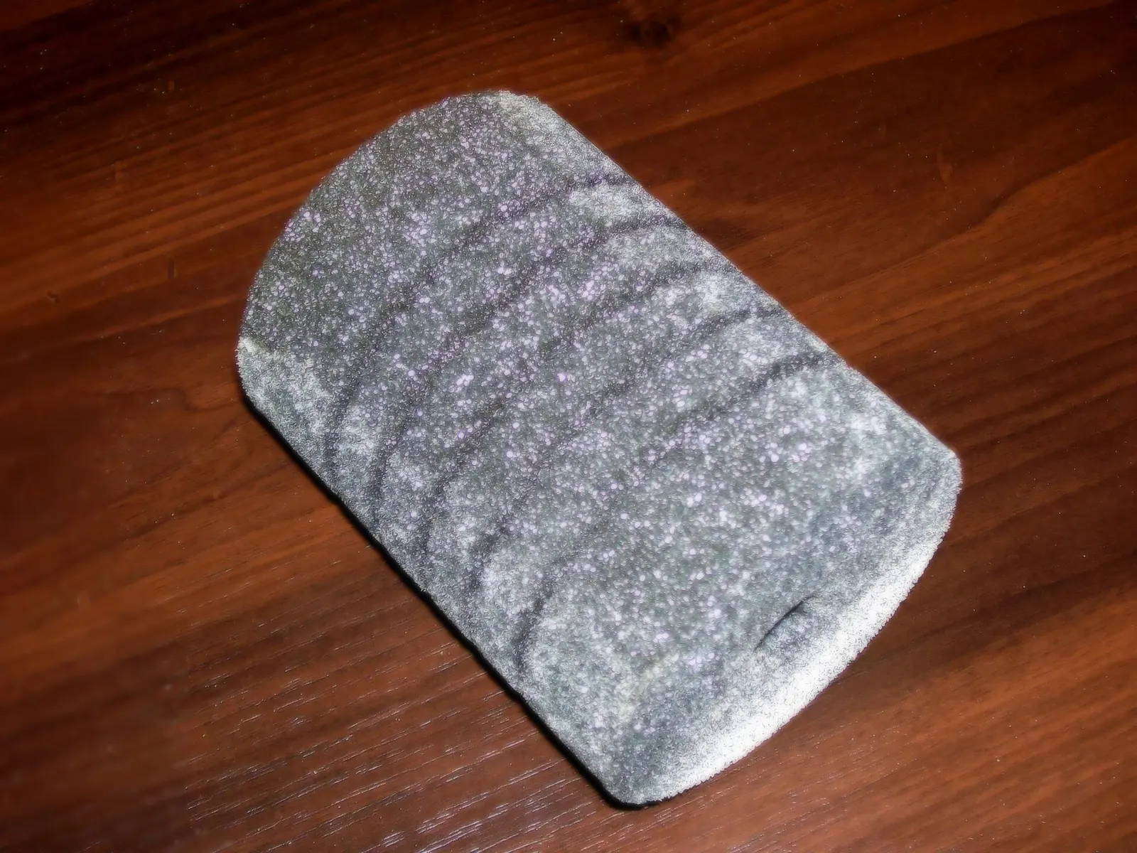 A used grey filter sponge with dark bands of trapped debris lying on a wooden surface