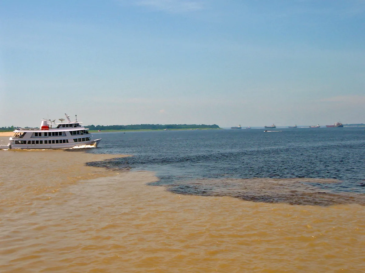 Dark Rio Negro water meeting pale brown, silt-laden Solimões water near Manaus, with river boats on the dark side