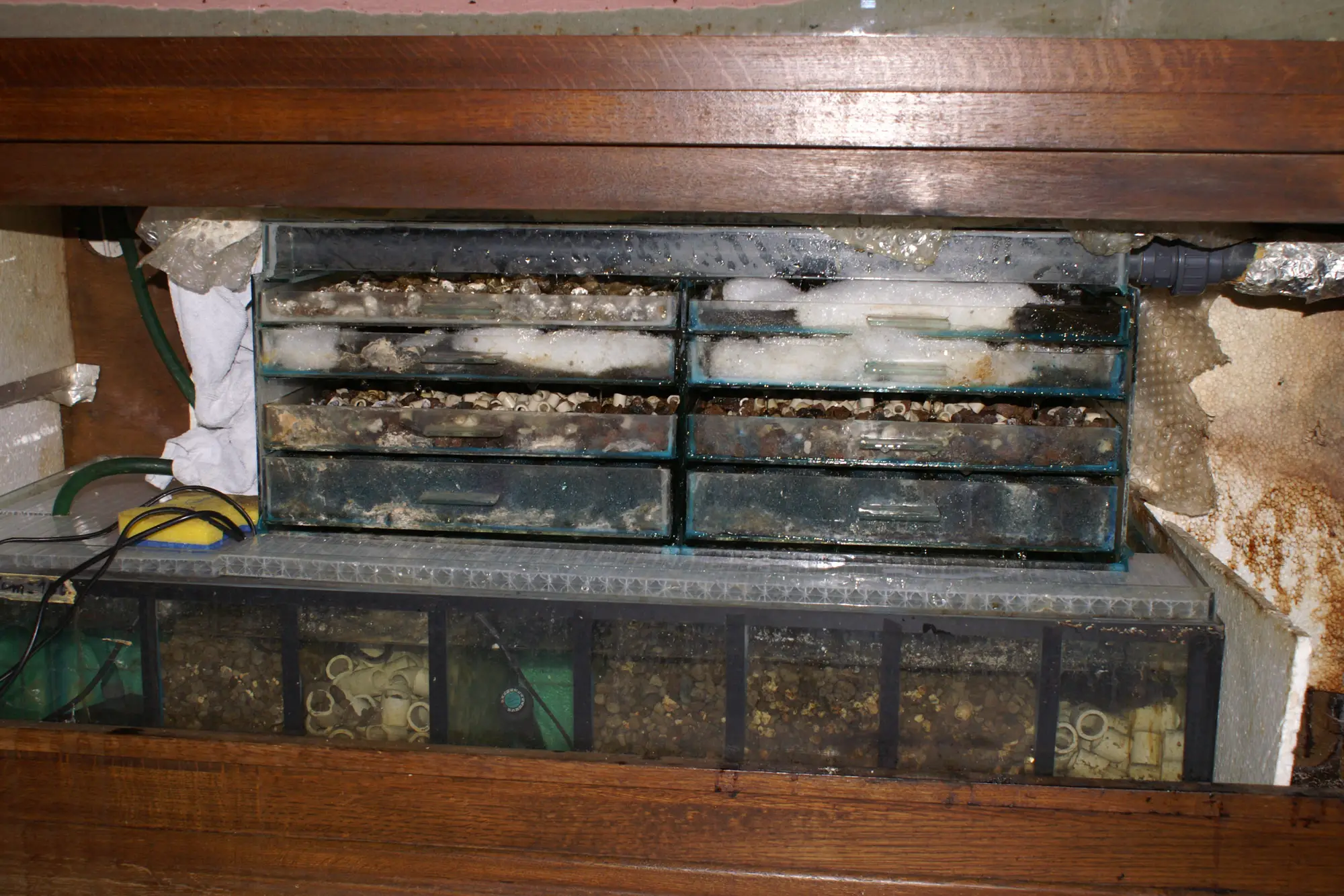 Trays of an established aquarium trickle filter packed with brown, coated ceramic rings above a sump of submerged media