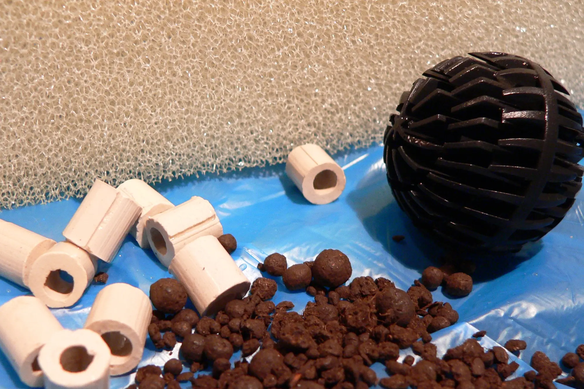 Unused aquarium filter media laid out on a blue sheet: pale ceramic rings, clay pebbles, a black plastic bio-ball and grey foam